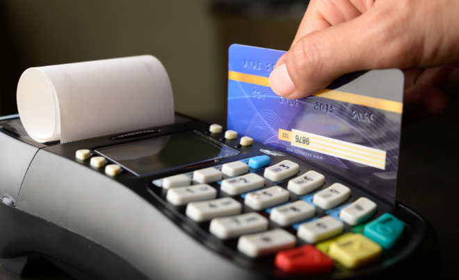 Tay đang quẹt thẻ ngân hàng qua máy POS, bên cạnh là cuộn giấy in hóa đơn.