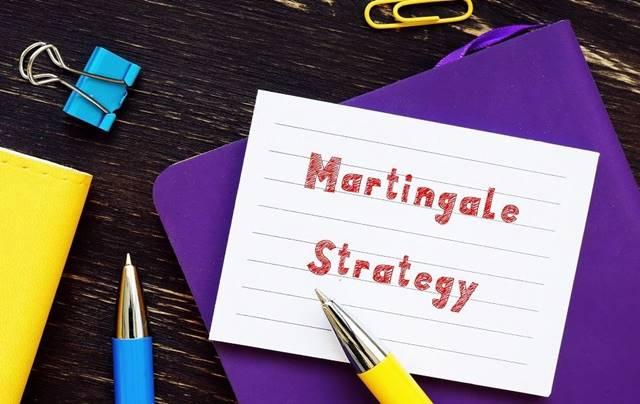 Tấm bìa giấy ghi "Martingale Strategy" đặt trên bàn, xung quanh có bút và kẹp giấy.