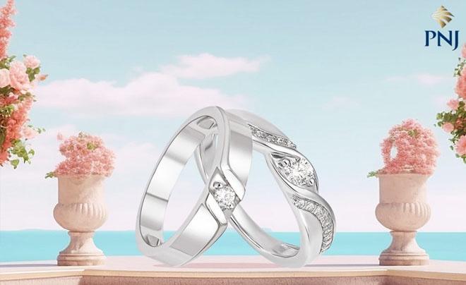 Hai chiếc nhẫn bạc có đính đá, đặt trên nền cảnh biển và trời xanh, xung quanh là các chậu hoa trang trí.