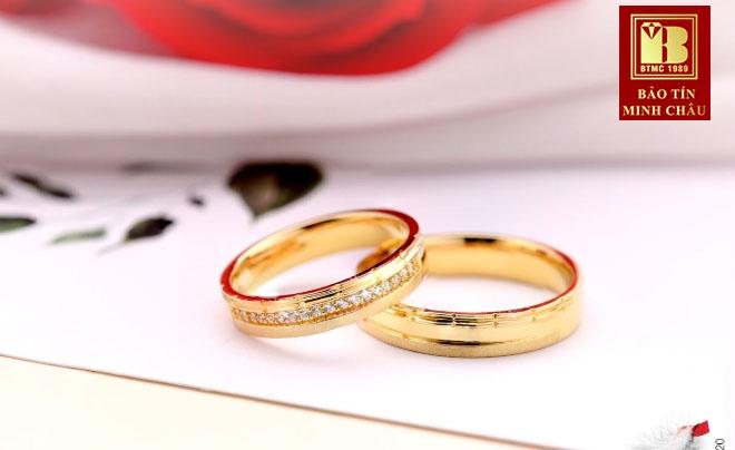 Hai chiếc nhẫn vàng được đặt trên nền trắng, phía sau là hình ảnh mờ của hoa hồng và logo thương hiệu.