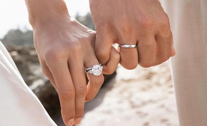 Hai bàn tay đeo nhẫn cưới nắm chặt nhau, nền là bãi biển và trời xanh.