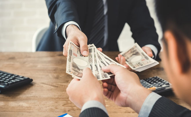 Hai người trao đổi tiền giấy trên bàn gỗ, có máy tính cầm tay và tài liệu xung quanh.