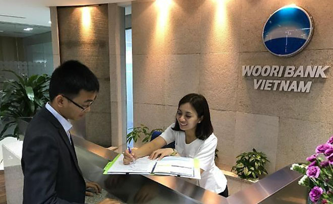 Người phụ nữ đang hỗ trợ khách hàng tại quầy giao dịch ngân hàng, với biển hiệu Woori Bank Vietnam phía sau.