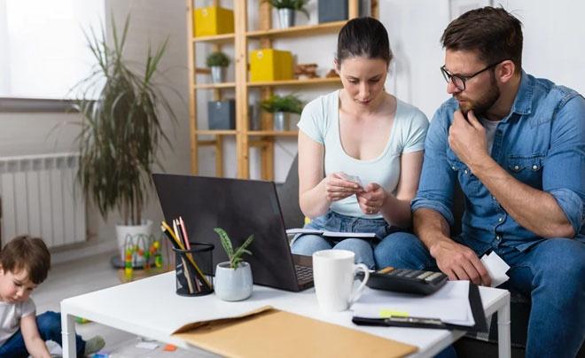Hai người ngồi bàn, xem tài liệu, với máy tính và máy tính cầm tay. Trẻ nhỏ chơi trên sàn cạnh đó.