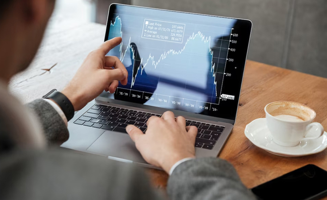 Người ngồi làm việc trên laptop với biểu đồ tài chính trên màn hình, bên cạnh là tách cà phê trên bàn gỗ.