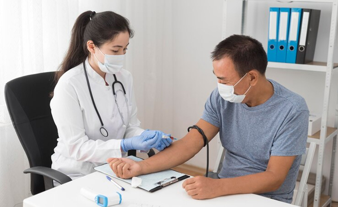 Nhân viên y tế đang đo huyết áp cho một bệnh nhân trong phòng khám, cả hai đều đeo khẩu trang.