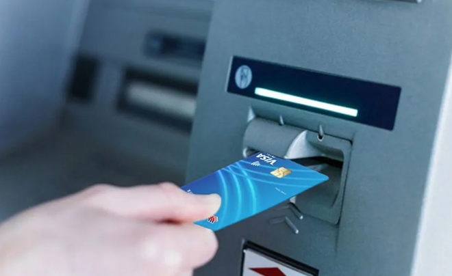 Tay đưa thẻ ngân hàng vào khe máy ATM với màn hình đèn sáng phía trên.