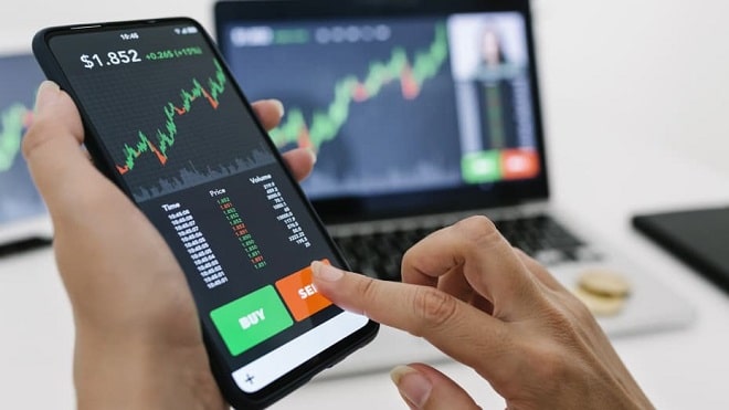 Người dùng thao tác trên ứng dụng giao dịch chứng khoán trên điện thoại, với biểu đồ giá hiển thị trên máy tính xách tay.