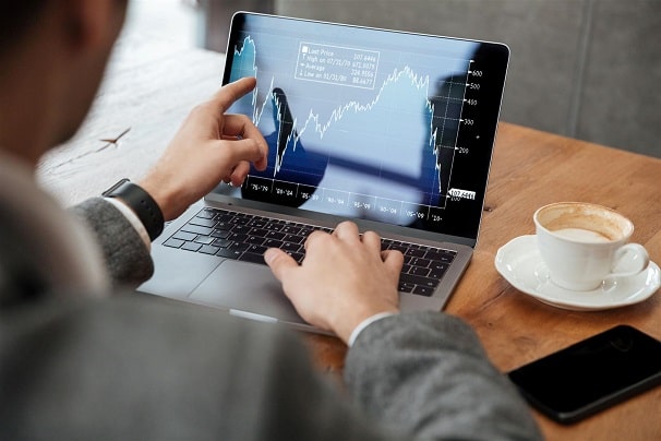 Người đang xem đồ thị tài chính trên màn hình laptop tại bàn làm việc có ly cà phê và điện thoại.
