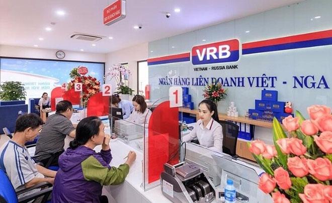 Ngân hàng với nhân viên đang giao dịch cùng khách hàng tại quầy dịch vụ, trang trí hoa và bảng hiệu ngân hàng.