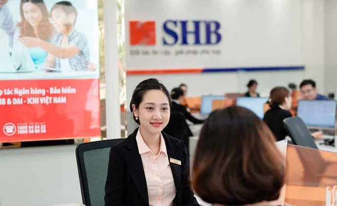 Nhân viên ngân hàng đang trò chuyện với khách hàng tại quầy giao dịch, nền có logo và bảng quảng cáo.