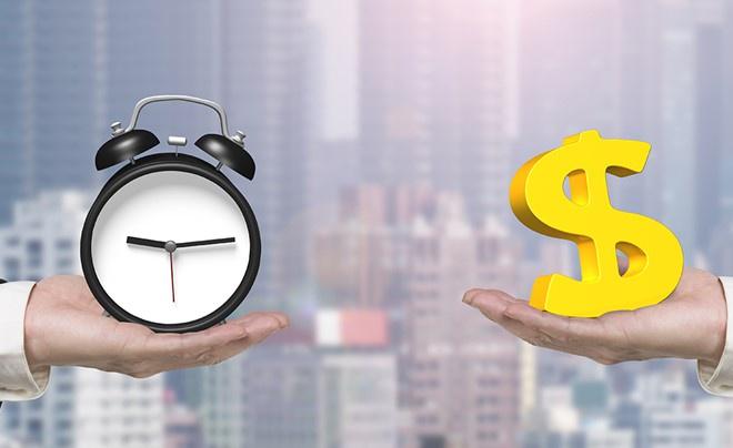 Hai tay cầm đồng hồ báo thức và biểu tượng đô la vàng, nền là tòa nhà mờ.