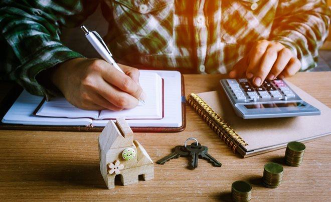 Người đang viết vào sổ tay và sử dụng máy tính, bên cạnh là mô hình ngôi nhà, chìa khóa và tiền xu.