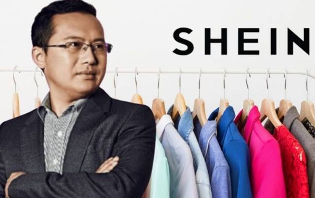 Người đàn ông đứng cạnh giá treo quần áo nhiều màu sắc, phía sau có chữ "SHEIN".
