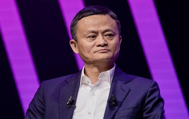 Một người đàn ông mặc vest xanh và áo sơ mi trắng ngồi trước nền màu tím.