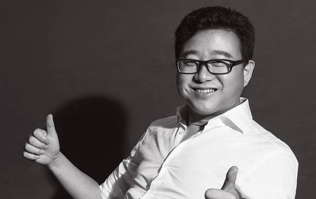 Người đàn ông đeo kính, mặc áo sơ mi trắng, cười và giơ ngón tay cái lên trong bức ảnh đen trắng.
