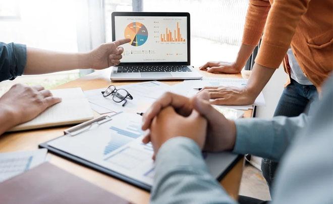 Nhóm người đang thảo luận trước máy tính xách tay hiển thị biểu đồ phân tích dữ liệu và tài liệu trên bàn.