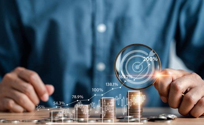 Người cầm kính lúp nhìn vào biểu đồ tăng trưởng tài chính với cột tiền xu và các chỉ số phần trăm.