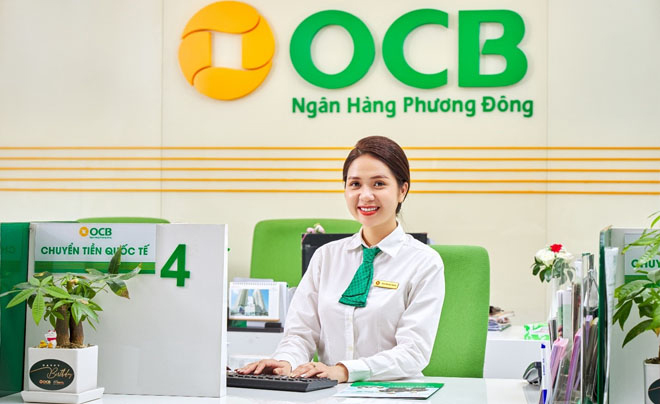 Nhân viên ngân hàng ngồi tại quầy giao dịch, mặc đồng phục trắng, nền có logo và tên ngân hàng.