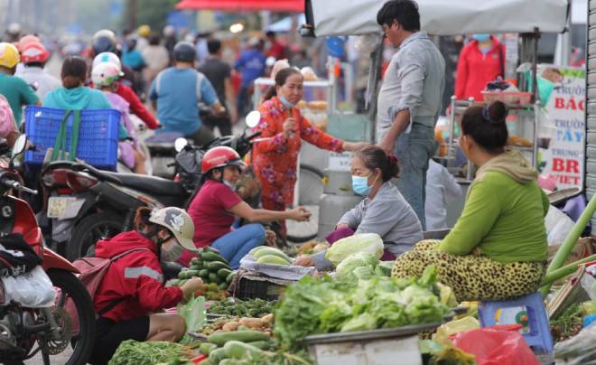 Chợ ngoài trời đông đúc, nhiều người mua bán rau củ tươi. Một số người ngồi trên xe máy, người khác đứng trò chuyện.