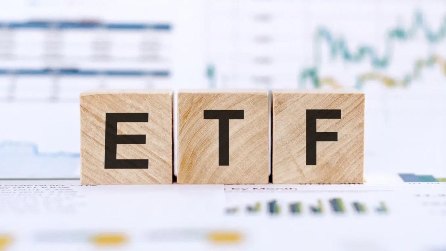 Ba khối gỗ có chữ "ETF" trên bề mặt, đặt trên nền biểu đồ tài chính mờ.