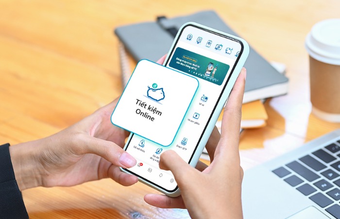 Người đang sử dụng ứng dụng tiết kiệm trực tuyến trên điện thoại thông minh, bên cạnh là laptop và cốc giấy.