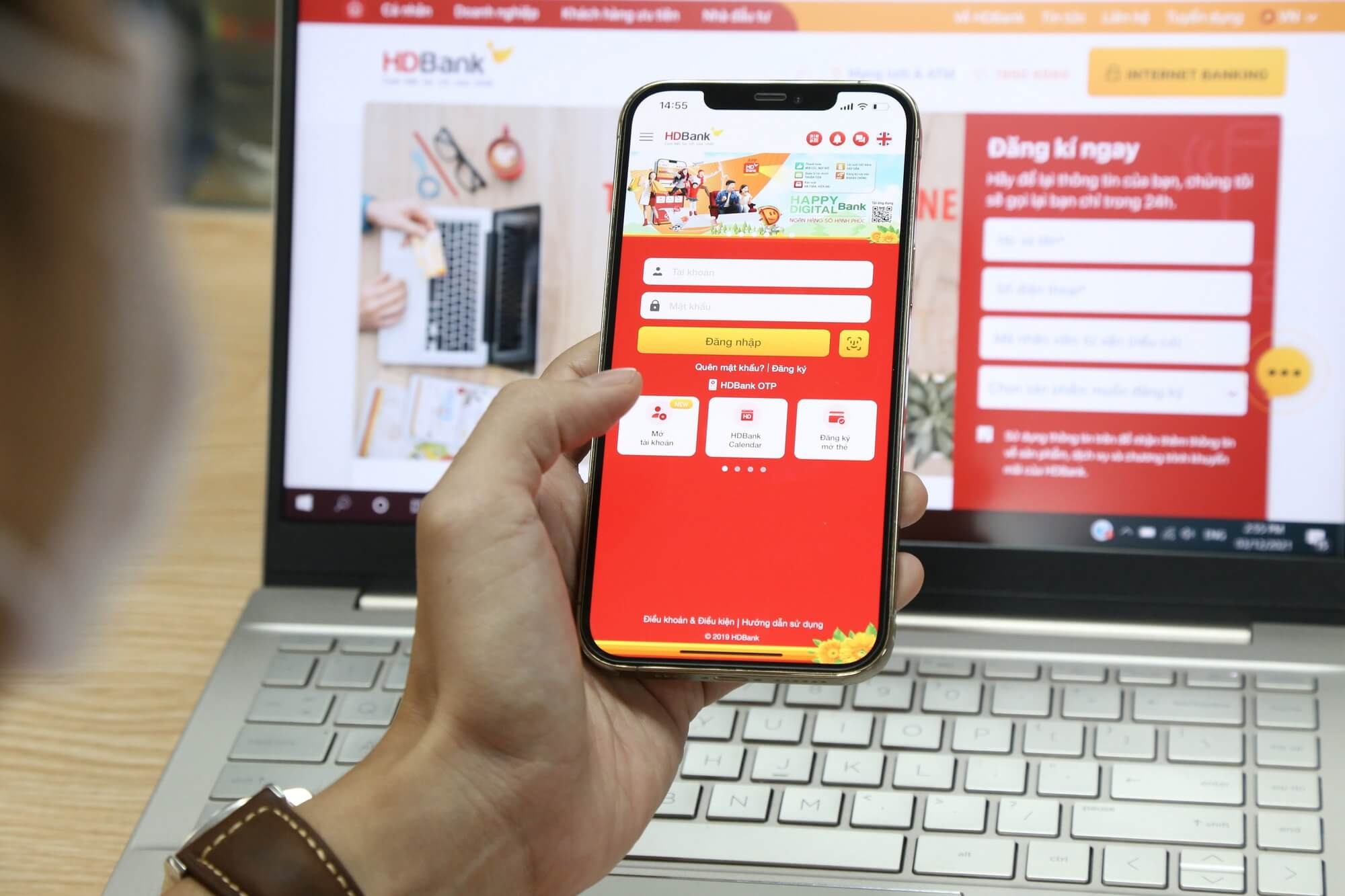 Người dùng đang đăng nhập ứng dụng ngân hàng trên điện thoại trước màn hình máy tính mở trang web ngân hàng.