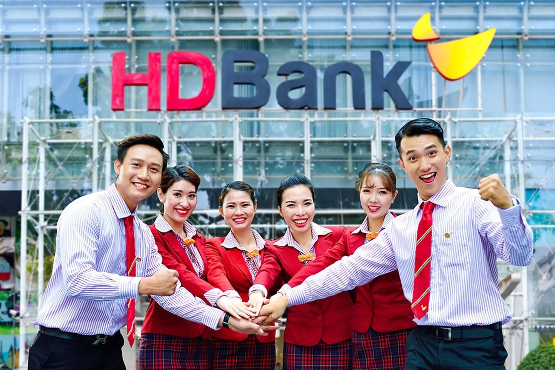 Nhóm người mặc đồng phục đỏ tươi cười, đứng trước tòa nhà có logo HDBank, tay chồng lên nhau thể hiện sự đoàn kết.