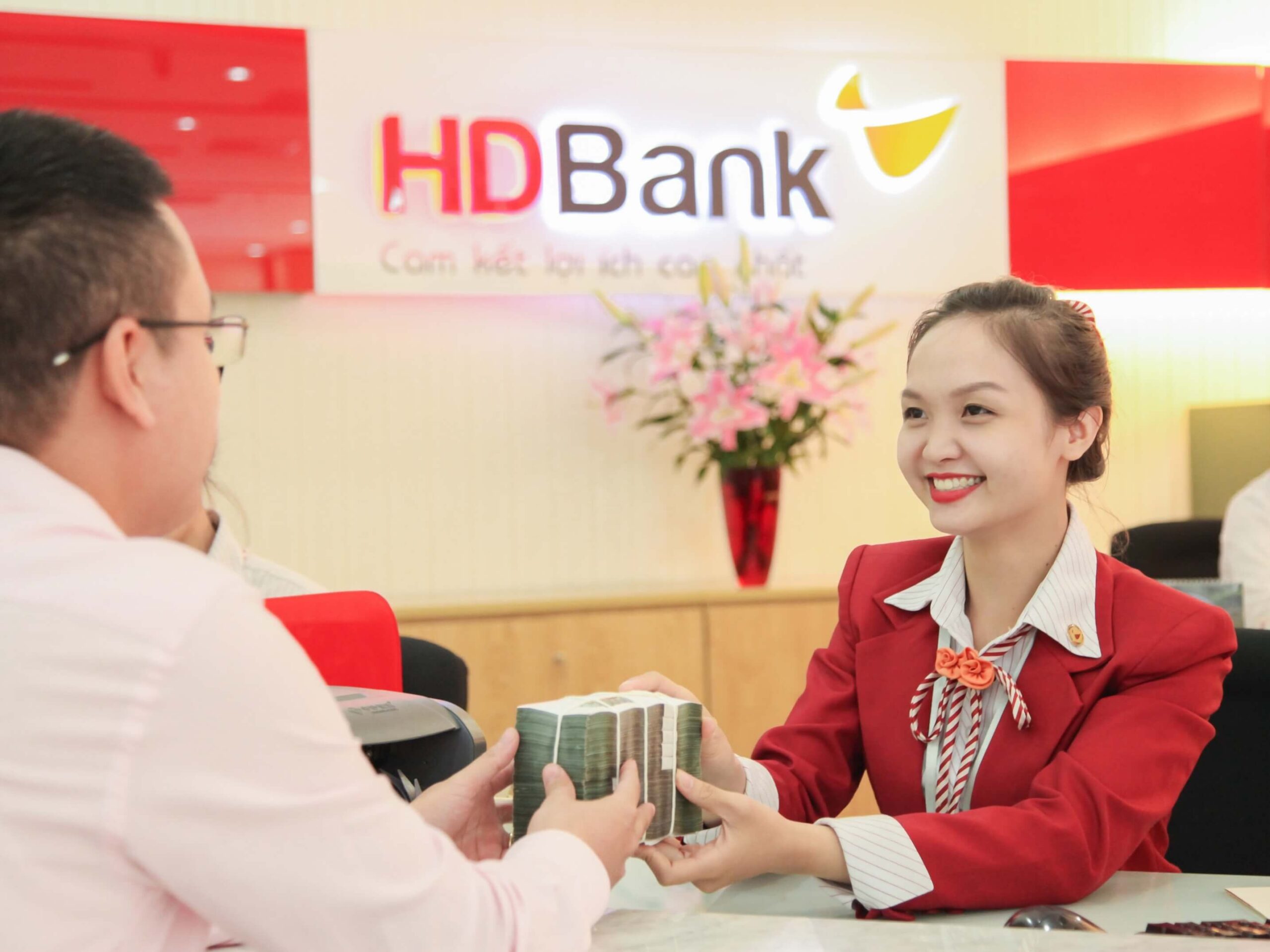 Nhân viên ngân hàng trao đổi tiền mặt với khách hàng tại quầy giao dịch, nền có logo ngân hàng.