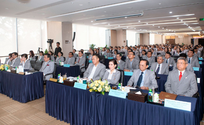 Hội trường đông người tham dự sự kiện, mọi người mặc trang phục trang trọng ngồi lắng nghe.