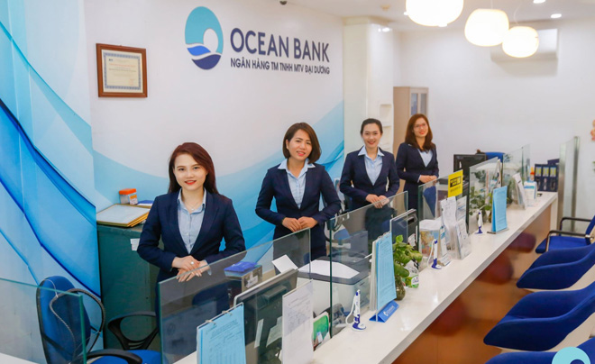 Nhân viên trong trang phục công sở đứng tại quầy giao dịch của một ngân hàng, với bảng hiệu "Ocean Bank" phía sau.