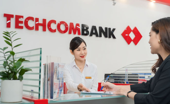 Nhân viên ngân hàng đang trao đổi với khách hàng tại quầy giao dịch, phía sau là logo của Techcombank.