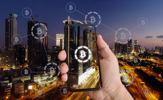 Người cầm điện thoại thông minh trên nền thành phố ban đêm với biểu tượng Bitcoin nổi trên bầu trời.