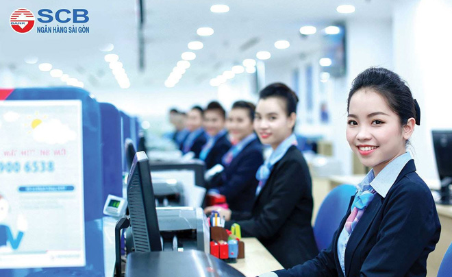 Nhân viên ngân hàng ngồi tại quầy giao dịch, mặc đồng phục, mỉm cười, trong không gian văn phòng sáng sủa.