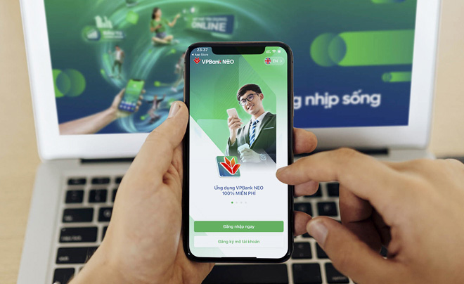 Người dùng cầm điện thoại hiển thị ứng dụng ngân hàng số trên nền laptop mở trang web liên quan.