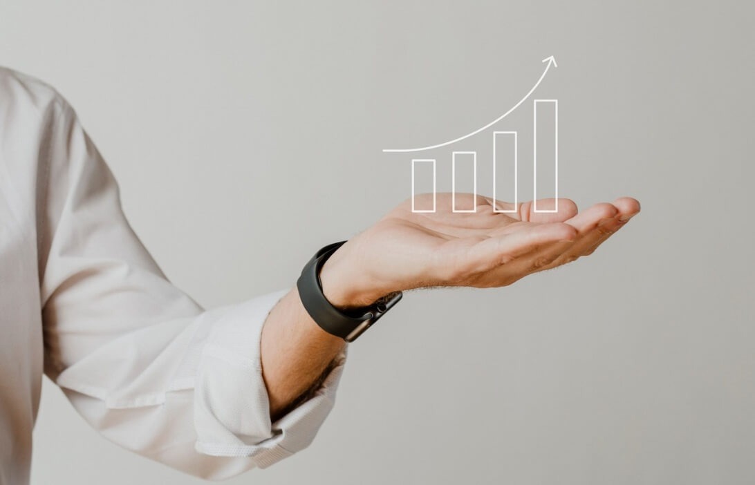 Bàn tay đang nâng một biểu đồ cột tăng trưởng với mũi tên hướng lên trên nền màu xám.
