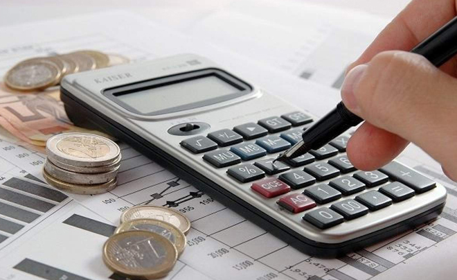 Một tay cầm bút nhấn máy tính trên bàn, cạnh đó là các biểu đồ tài chính và tiền xu.