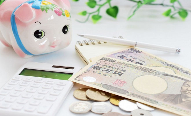 Ống heo, tiền giấy và tiền xu Nhật Bản, máy tính và sổ tay với bút trên bàn trắng, nền có lá cây xanh.