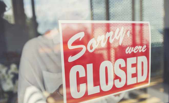 Tấm biển "Sorry, we're closed" treo trên cửa kính, phản chiếu hình bóng mờ nhạt của một người phía sau.