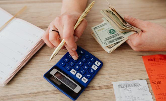 Người đang sử dụng máy tính và cầm tiền mặt, cạnh đó là sổ ghi chép và hóa đơn trên bàn gỗ.
