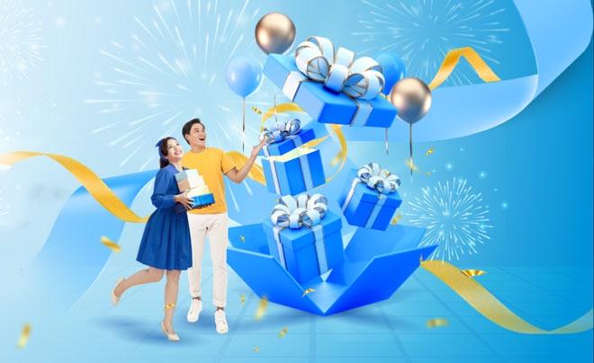 Hai người vui vẻ đứng cạnh hộp quà xanh lớn, xung quanh là pháo hoa và ruy băng vàng.