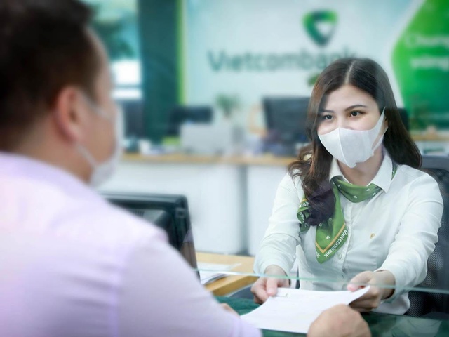 Nhân viên ngân hàng đeo khẩu trang đang trao đổi với khách hàng qua tấm kính tại quầy giao dịch.