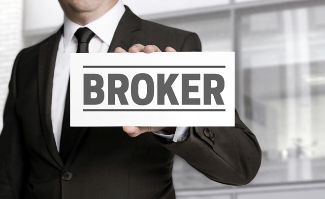 Người mặc vest cầm tấm bảng có chữ "BROKER" trên nền mờ của tòa nhà văn phòng.