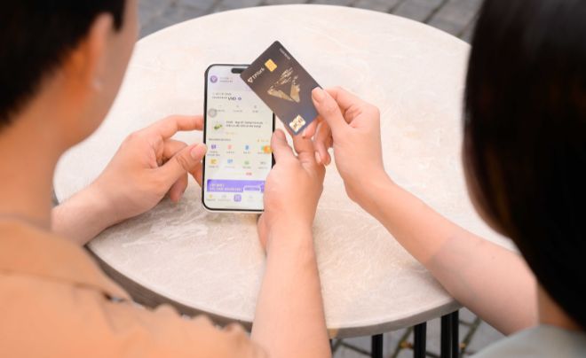 Hai người ngồi tại bàn, một người cầm điện thoại, người kia cầm thẻ ngân hàng.