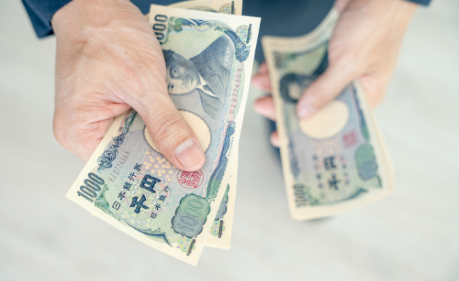 Đôi tay đang cầm tiền giấy Nhật Bản, mệnh giá 1.000 yên, trên nền mờ.