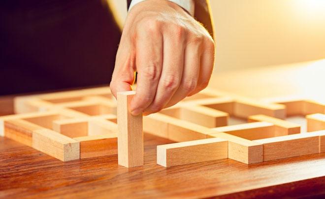 Bàn tay đang đặt một khối gỗ đứng vào mô hình mê cung bằng gỗ trên mặt bàn gỗ dưới ánh sáng vàng.