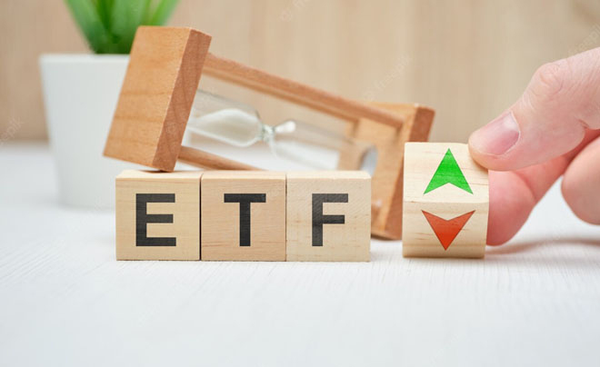Khối gỗ chữ ETF, biểu tượng mũi tên lên xuống và đồng hồ cát trên bàn trắng.