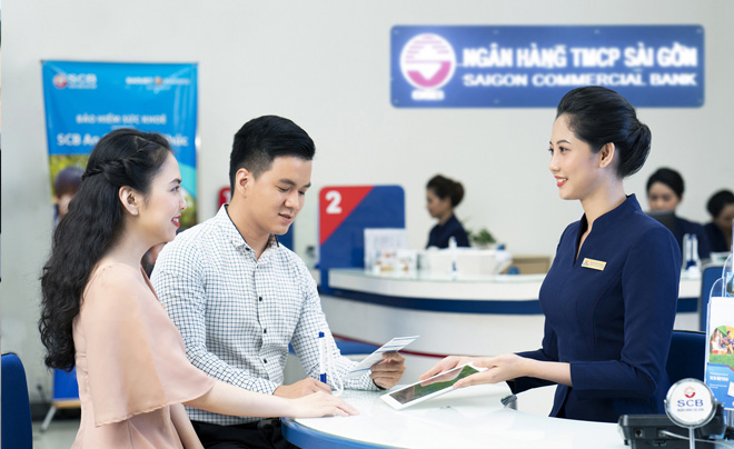 Nhân viên ngân hàng trao đổi với khách hàng tại quầy giao dịch, nền có biển hiệu ngân hàng.