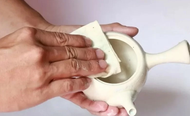 Hai tay đang lau chiếc ấm trà bằng một miếng vải trắng.