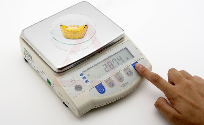 Cân điện tử đo trọng lượng một vật nhỏ màu vàng, hiển thị 28.74g, với một ngón tay đang nhấn nút trên màn hình.